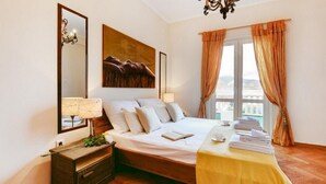 A hotel room with a large bed, a bedside table with a lamp, a painting on the wall, and a view of the sea through the window.