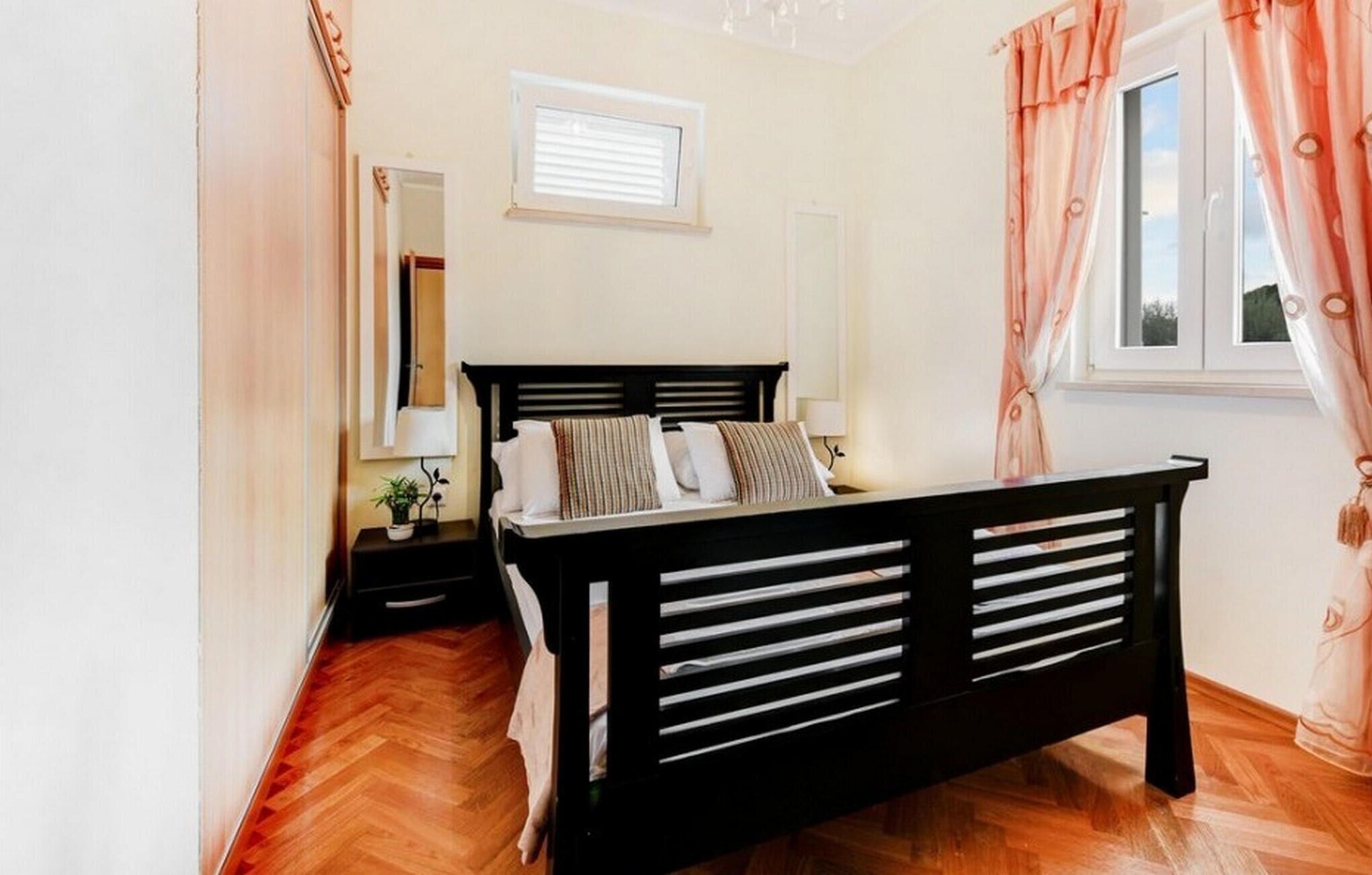 A bedroom with a wooden bed, a nightstand, a window with curtains, and a mirror on the wall.