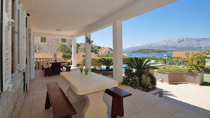 A covered outdoor area with a table and benches, a pool, and a view of the sea and mountains.