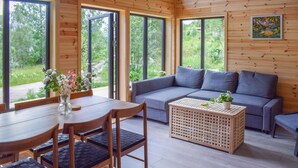Une pièce chaleureuse avec un décor intérieur en bois, un canapé gris, une table à manger avec des chaises et de grandes fenêtres donnant sur un paysage verdoyant.