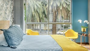 Ein modernes Schlafzimmer mit einem großen Fenster, einem Bett mit blau-gelber Bettwäsche, einem gelben Sessel und einem kleinen Tisch mit Pflanzen.
