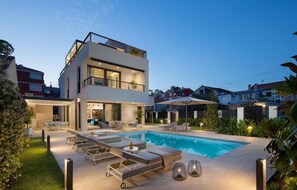 A modern two-story house with a swimming pool, outdoor seating, and a well-lit patio area.