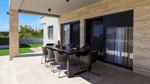 A modern outdoor dining area with a table and chairs, large sliding glass doors, and a view of a green lawn.