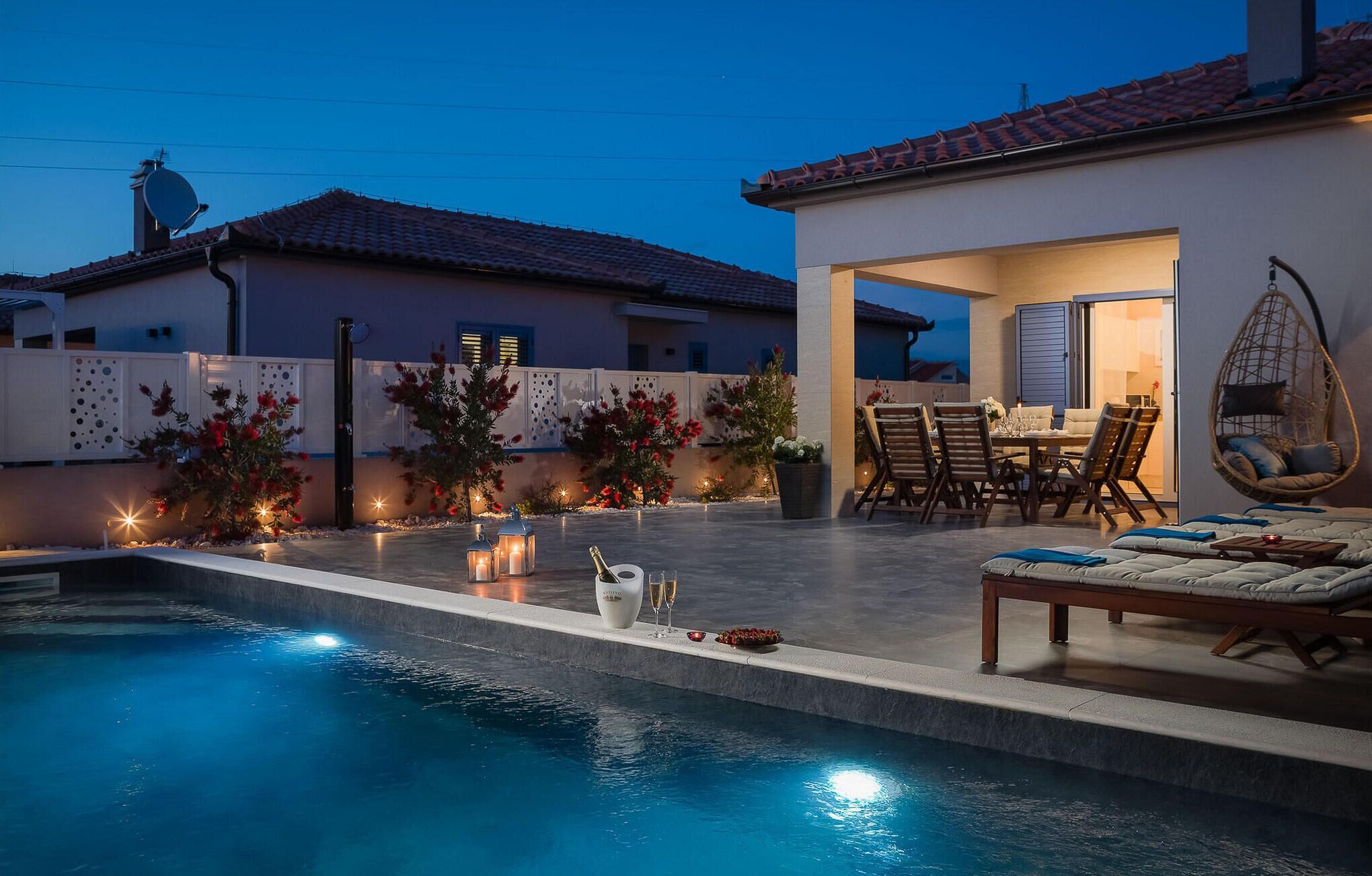 A swimming pool with a patio area, outdoor furniture, and a view of a house with a tiled roof.