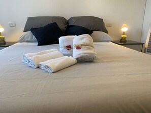Een netjes opgemaakt bed met witte en grijze lakens, twee witte handdoeken met rode details, en twee zwarte kussens.