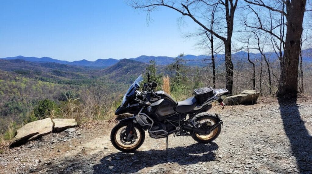 A motorcycle parked on a gravel path with a scenic view of mountains and trees in the background.