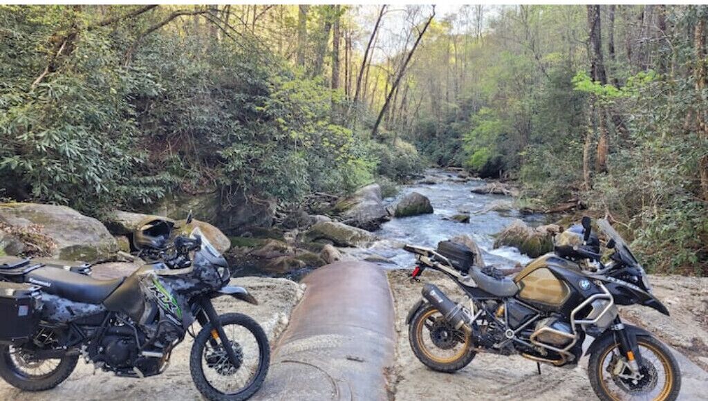 Two motorcycles parked on a rocky path beside a flowing river in a forested area.