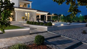 Une maison moderne avec une façade en pierre, un patio couvert aménagé avec du mobilier d’extérieur et un jardin bien éclairé.