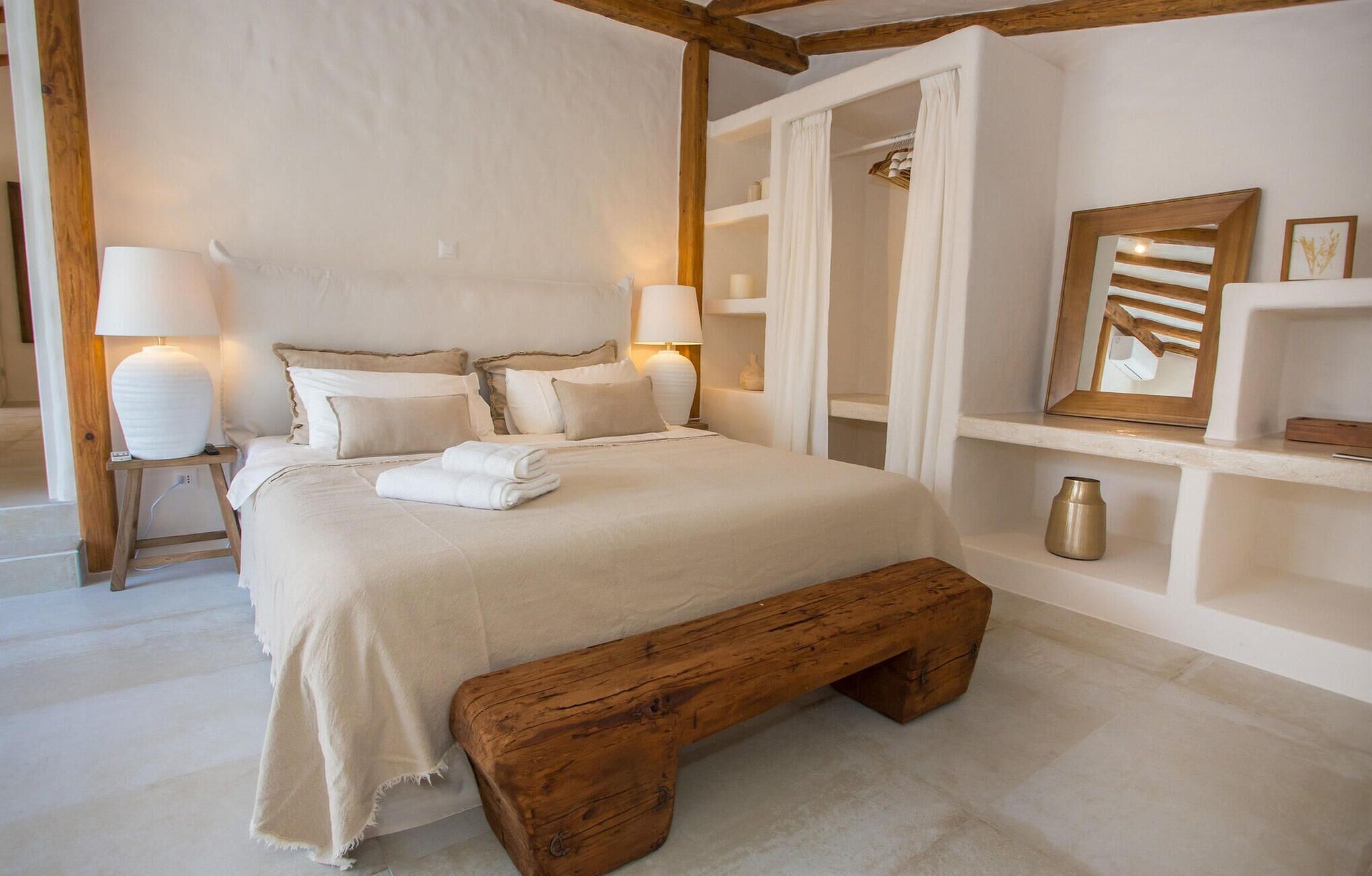 A neatly arranged bedroom with a large bed, bedside lamps, a wooden bench, and a mirror on the wall.