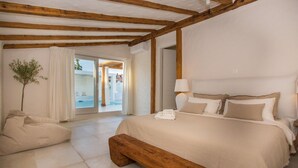 Ein Schlafzimmer mit einem großen Bett, einer Bank, einem Nachttisch, einer Lampe und Blick auf einen Außenpoolbereich.