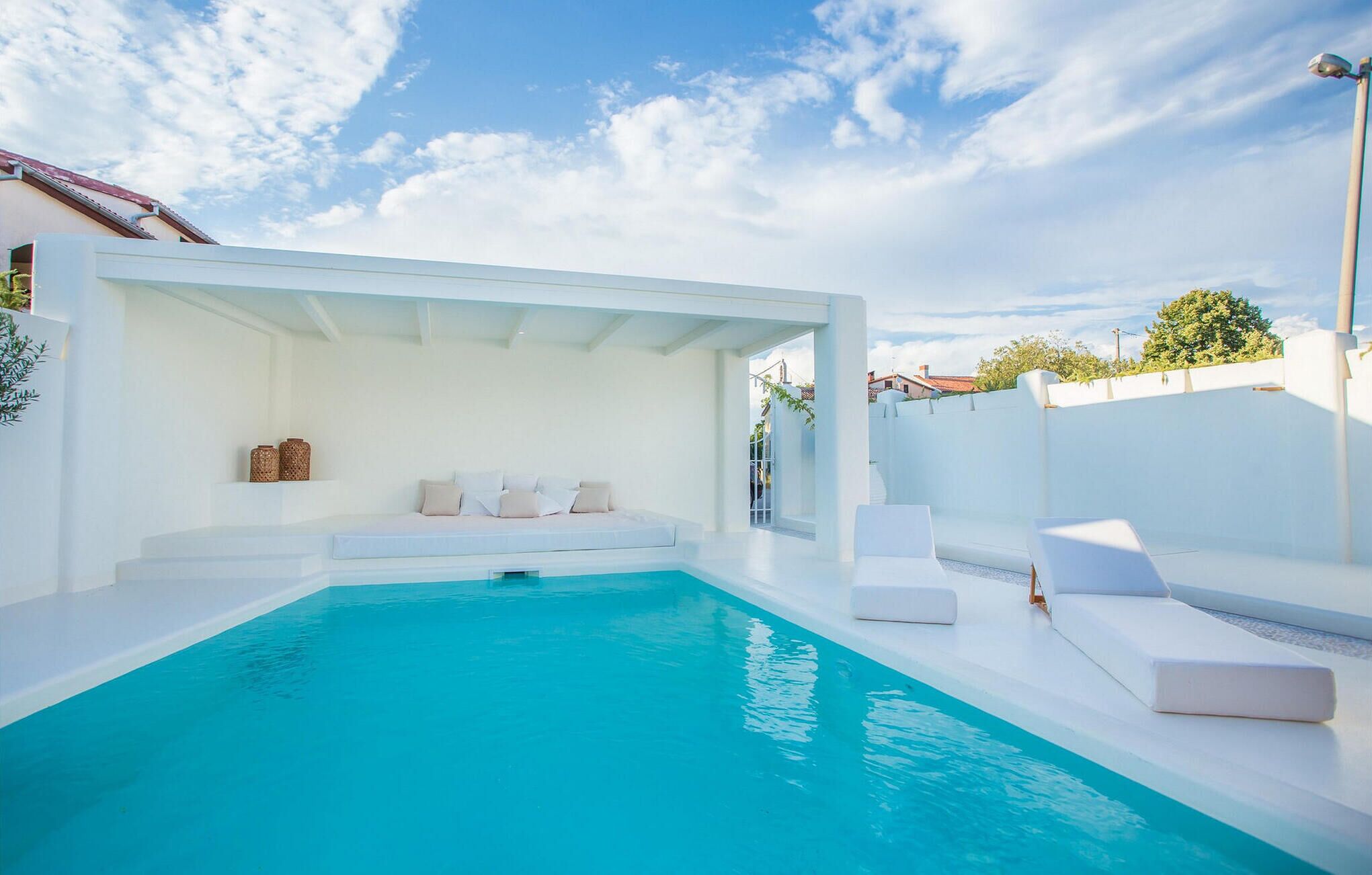 A modern outdoor pool area with white walls and furniture, a clear blue sky, and a view of the surrounding neighborhood.