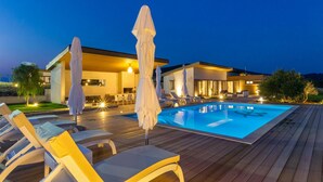 A modern outdoor pool area with lounge chairs and umbrellas, a wooden deck, and a contemporary house in the background. - 5 bedroom gorgeous home in Hvar (Hvar)