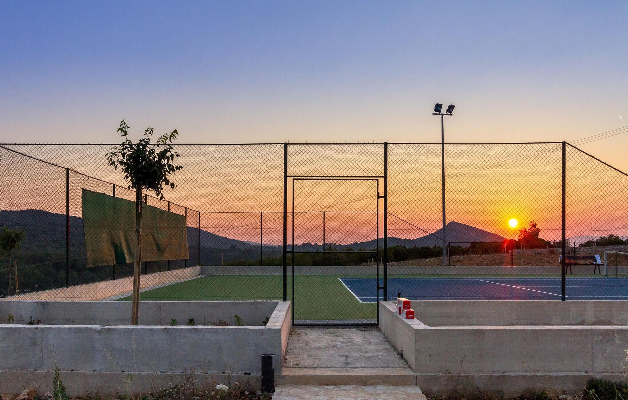 Un court de tennis avec un filet, entouré d’une clôture en treillis métallique, et un coucher de soleil en arrière-plan.