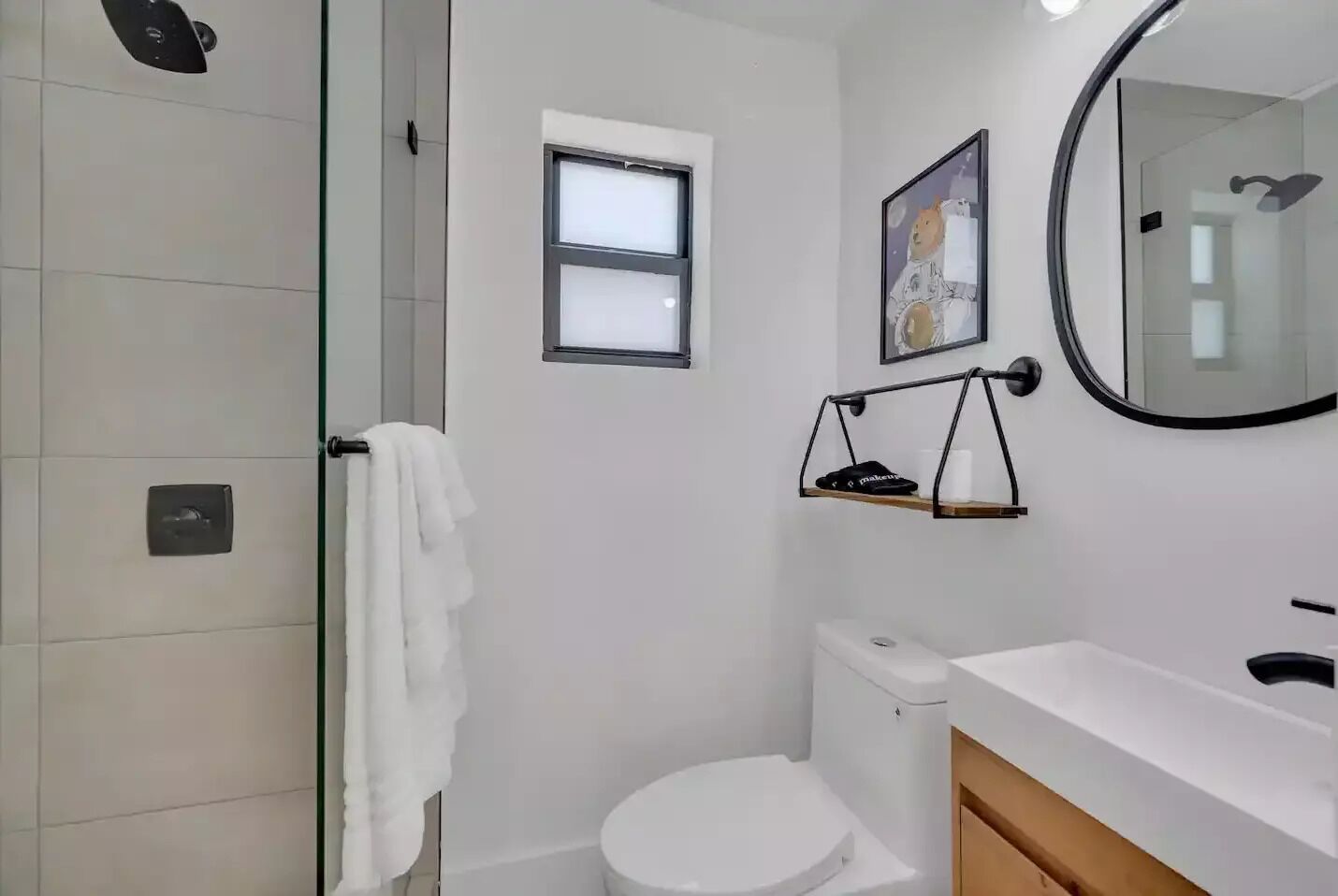 A modern bathroom with a glass shower enclosure, a white toilet, a small window, and a wall-mounted shelf with a towel.