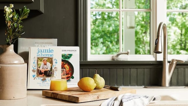 Une cuisine avec des meubles de couleur sombre, une fenêtre donnant sur un paysage verdoyant, un évier et un plan de travail sur lequel se trouvent un livre et des fruits.
