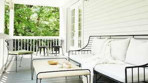 Une véranda avec une balustrade blanche, un banc en métal noir, un canapé blanc et une table sur laquelle se trouvent un saladier et des tasses.