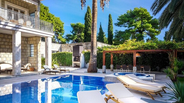 A pool area with lounge chairs, a pergola, and a stone building.