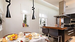 A modern kitchen with a dining area, featuring a breakfast setup with fruits, pastries, and tea.