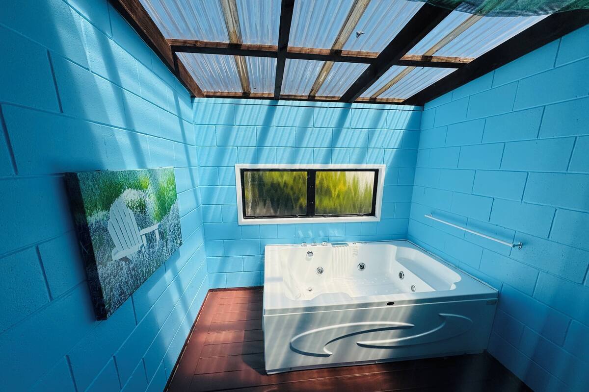 Un baño moderno con paredes azules, piso de madera, una bañera grande y una ventana con vistas a follaje.