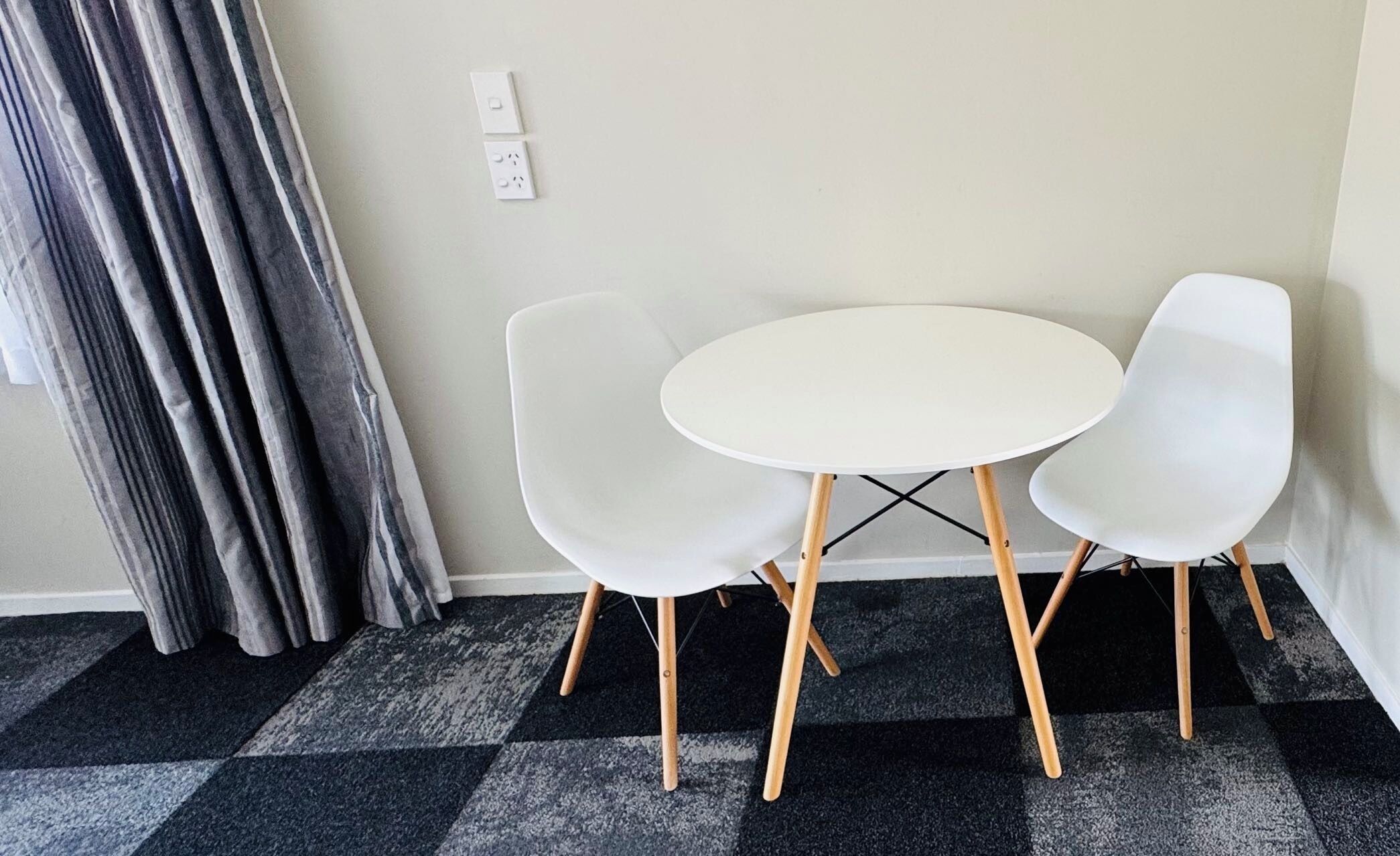 A room with a checkered carpet, a white round table, and two white chairs with wooden legs.