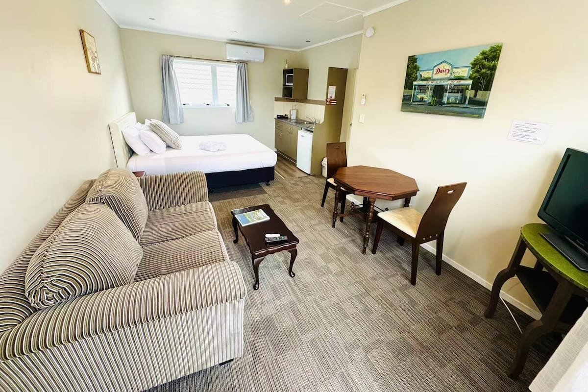 Habitación de hotel pequeña con cama, sofá, mesa de comedor con sillas, televisor pequeño y un cuadro en la pared.