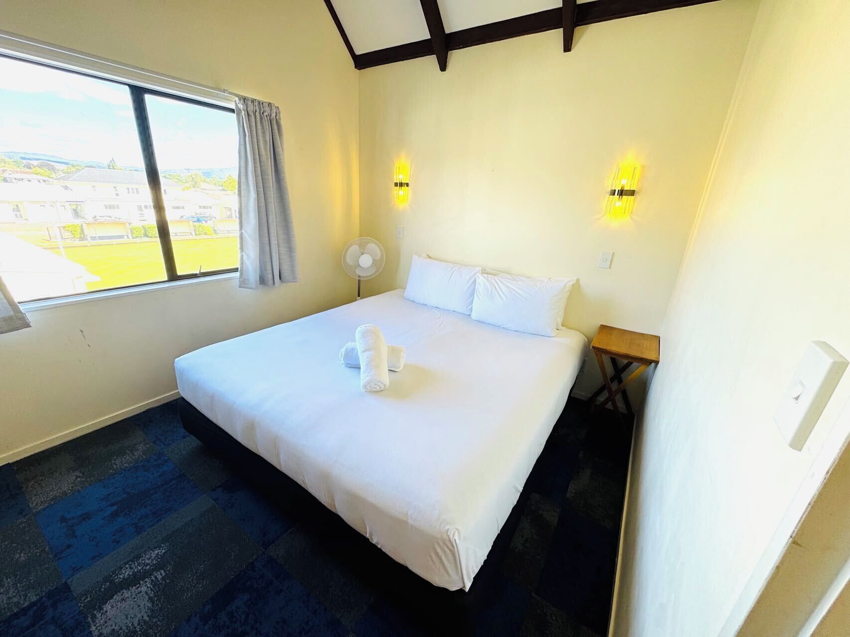 Ein Hotelzimmer mit einem großen Bett, zwei Nachttischlampen, einem fensterbelaubten Fenster, einem Standventilator und einem kleinen Holztisch.
