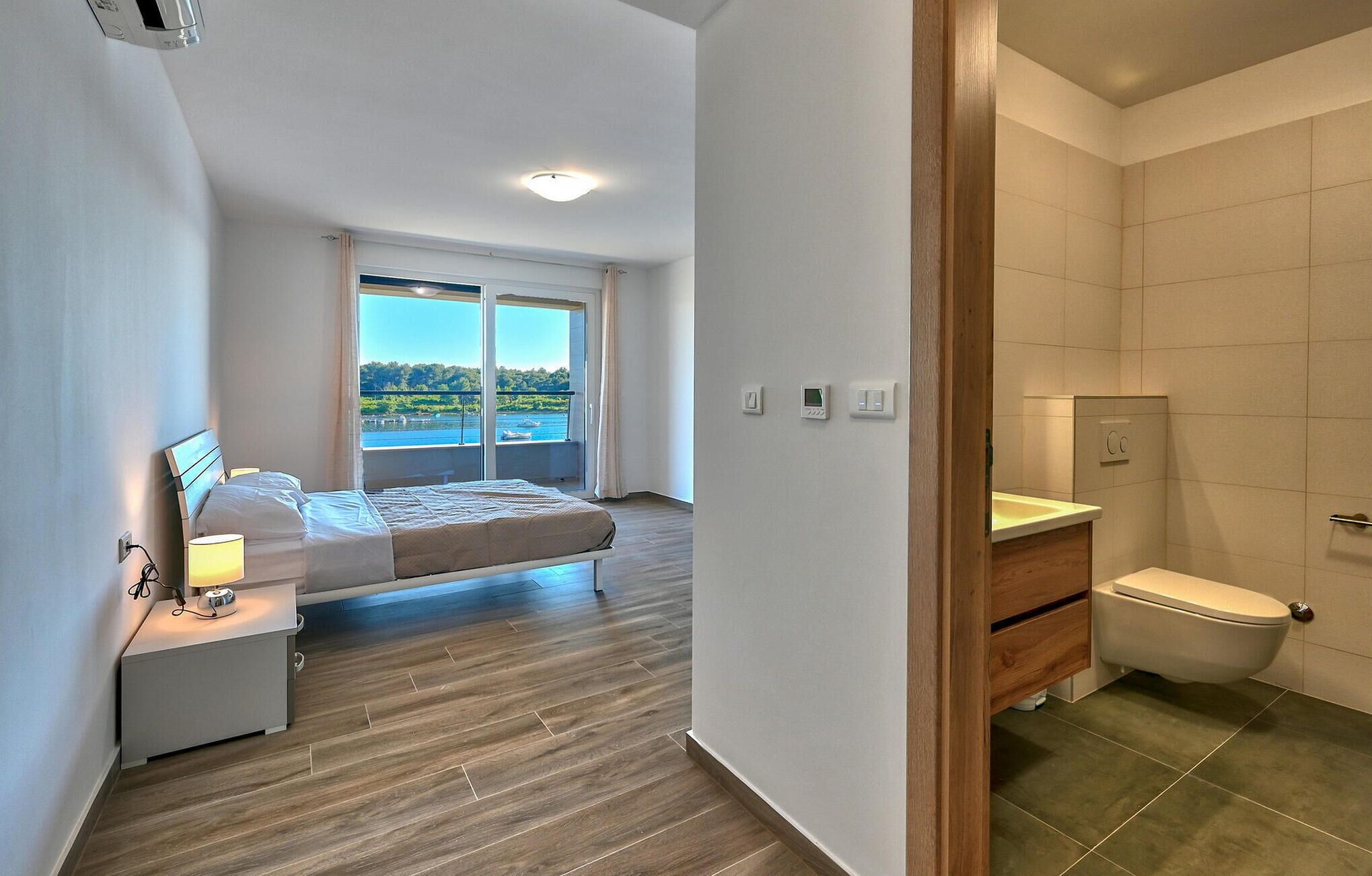 Un dormitorio con cama, lámparas de noche, ventana con vista al mar y baño con inodoro y lavamanos.