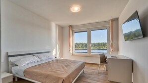 Ein Schlafzimmer mit einem Bett, einem Schreibtisch, einem Sessel, einem Fernseher und einem Fenster mit Blick auf einen See und Bäume.