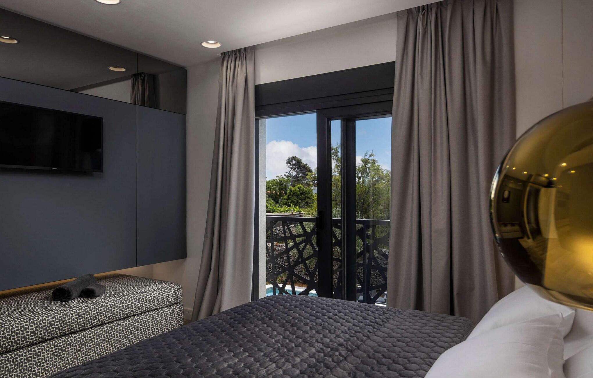 A modern hotel room with a large bed, a TV mounted on the wall, and a balcony with a view of greenery.