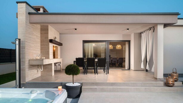 Een modern terras buiten, met een hottub, een eethoek en een glazen deur naar een binnenruimte.