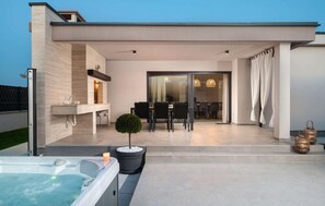 A modern outdoor patio with a hot tub, a dining area, and a glass door leading to an interior space.