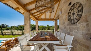 Un espacio exterior para comer con mesa de madera y sillas blancas, un muro de piedra con un reloj y vistas a una piscina y follaje.