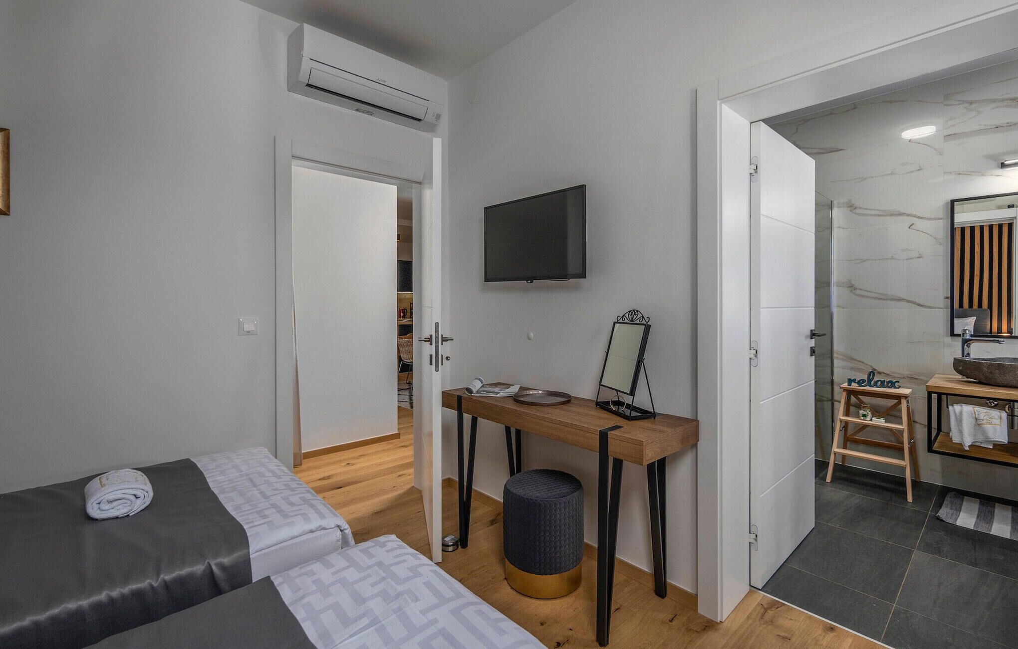 A modern hotel room with a bed, a desk with a mirror, a television, and a bathroom with a marble floor.