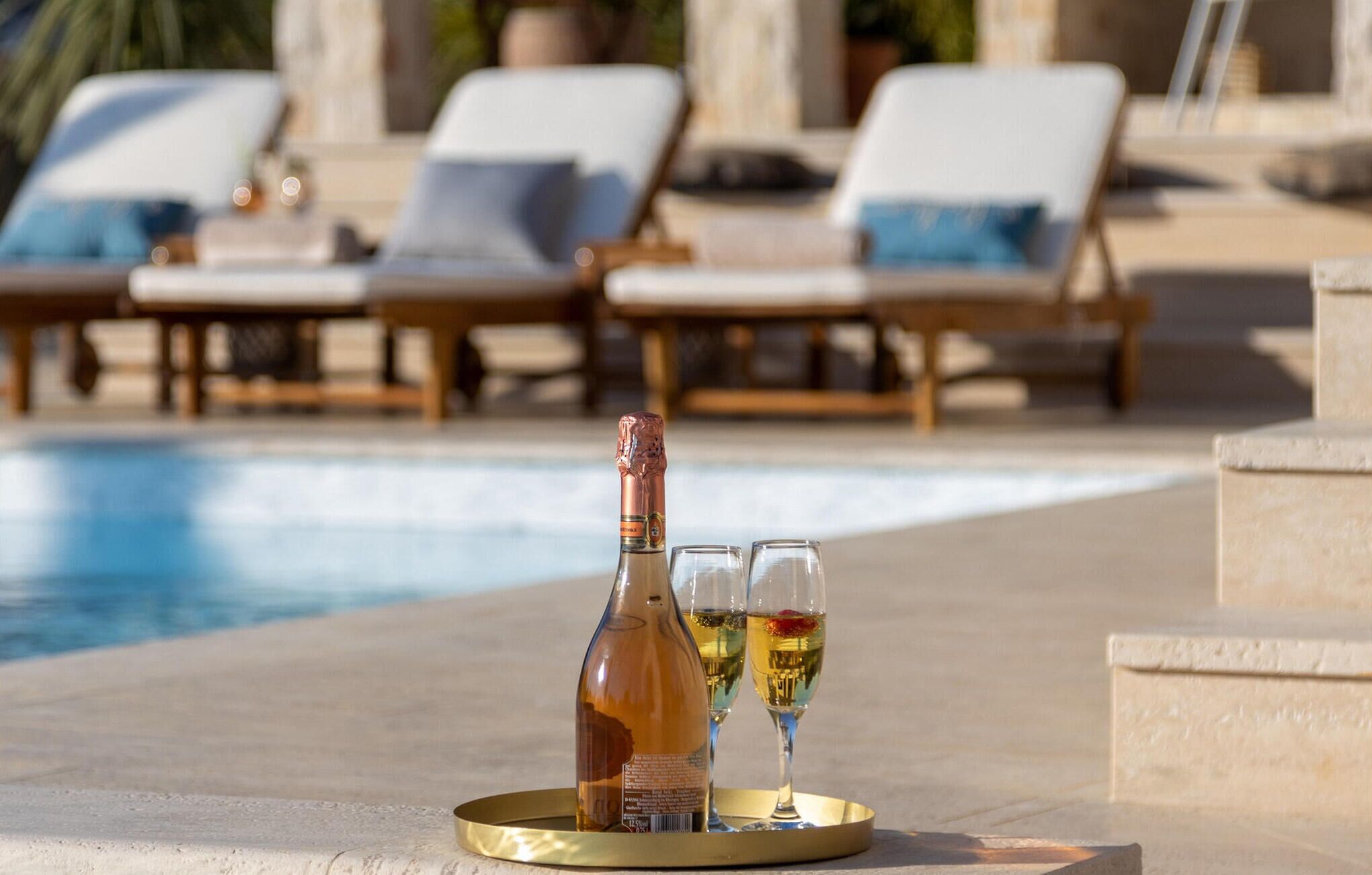 Une bouteille de champagne et deux verres sur un plateau, placés près d’une piscine, avec des chaises longues en arrière-plan.