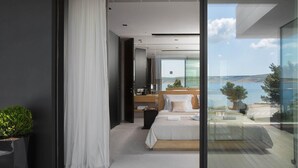 A modern bedroom with a large bed, a wooden headboard, and a view of the sea through floor-to-ceiling glass windows.