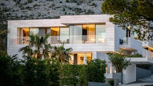 A modern multi-story building with large glass windows, a flat roof, and a landscaped garden with palm trees.