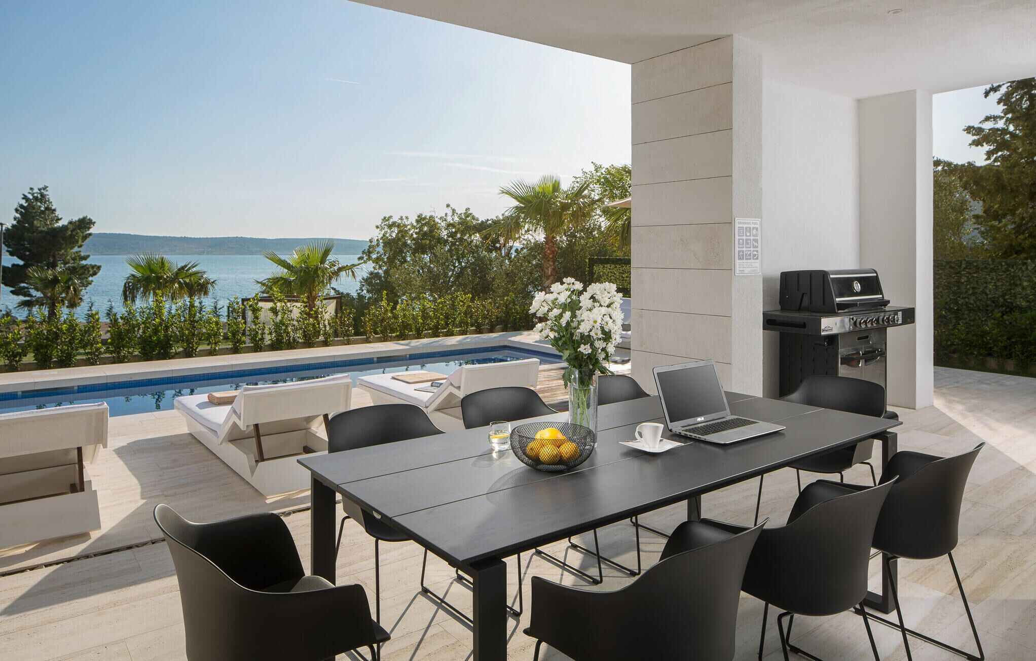 A modern outdoor dining area with a pool, a grill, and a view of the sea.