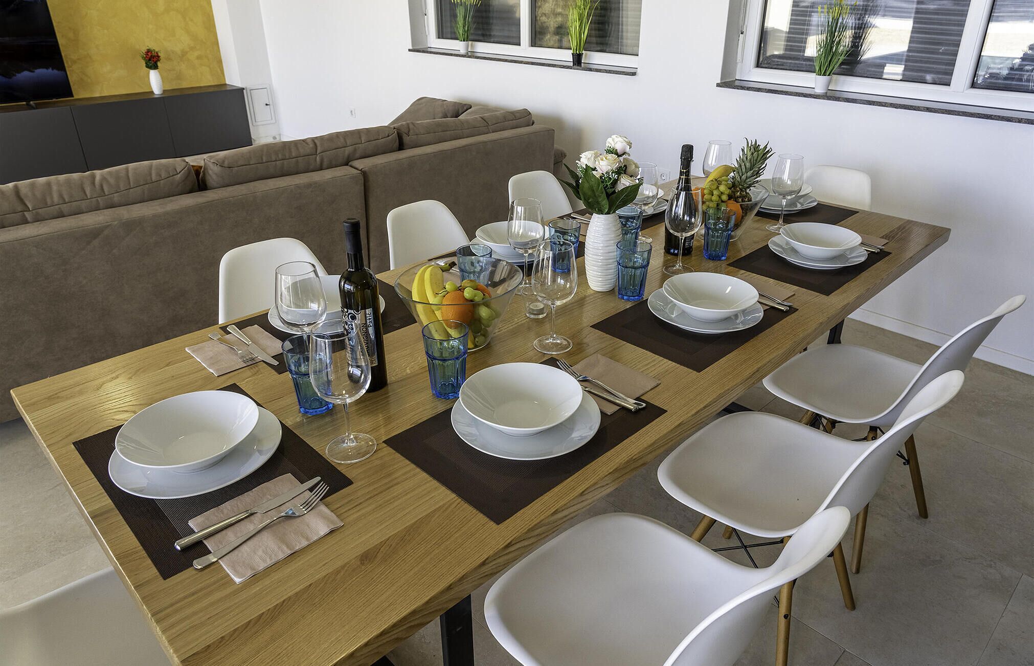 A dining table set with white plates, bowls, glasses, and cutlery, surrounded by white chairs.