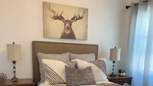 Ein Schlafzimmer mit einem großen Bett, zwei Nachttischen, einem Beistelltisch, einer dekorativen Lampe, einem Teppich und einem Bild von einem Reh an der Wand.