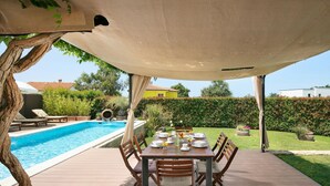 Ein Essbereich im Freien mit Pool, Holzterrasse und einem überdachten Tisch mit Stühlen.