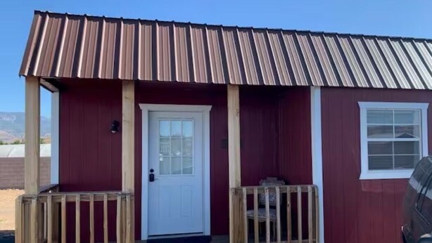 A small, single-story building with a red exterior and a brown metal roof. It features a front door with a small porch area. - The Wild West's Best Kept Secret  (Hurricane)