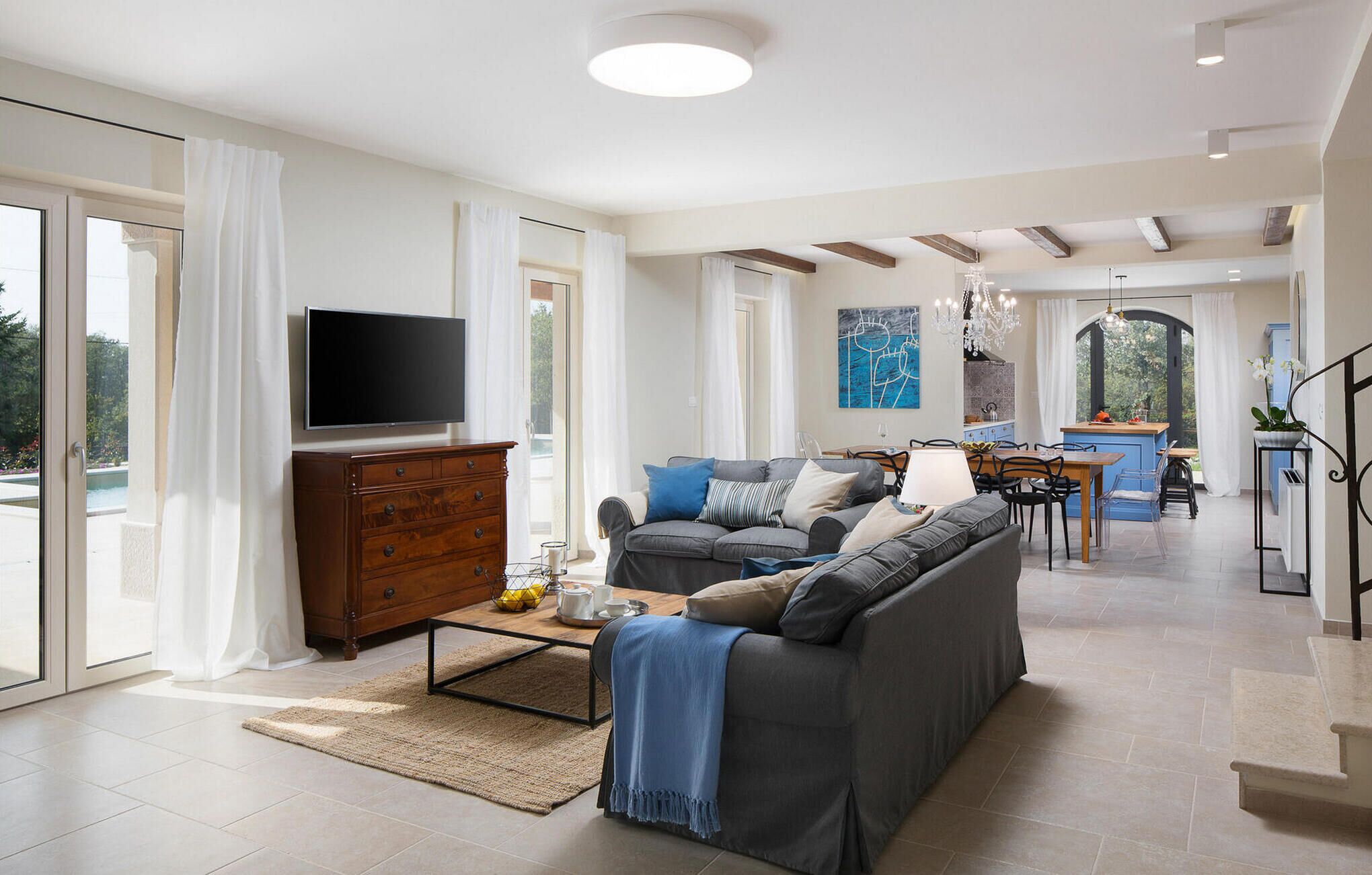 A spacious living area with a sofa, coffee table, and TV. There is a dining area with chairs and a table. The room has large windows and a view of the outdoors.