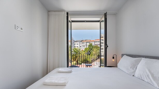 A hotel room with a large bed, white linens, a view of the city through an open door, and a balcony with a railing. - GuestReady - Serene stay in Porto (Porto)