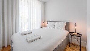 A modern hotel room with a large bed, white linens, a bedside table with a lamp, and sheer curtains. - GuestReady - Serene stay in Porto (Porto)