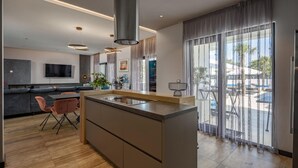 Une cuisine moderne dotée d’un îlot central, d’un coin repas avec des chaises, d’un téléviseur fixé au mur et de grandes fenêtres avec des rideaux.