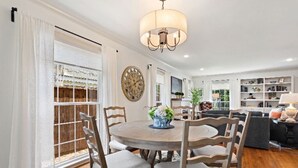 A dining area with a round table, chairs, and a chandelier. There is a large window with curtains and a view of a wooden structure outside.