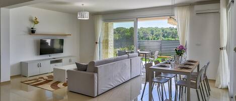 En moderne stue med et fladskærms-tv, en sofa, et spisebord med stole og et stort vindue med udsigt til grønne områder.