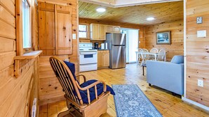 A wooden cabin interior with a rocking chair, a sofa, a dining table with chairs, a kitchen area with a refrigerator and stove, and a patterned rug.