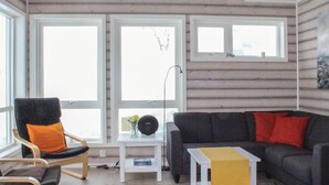 Ein gemütliches Blockhaus-Interieur mit Holzbalkendecke, einem schwarzen Sessel mit orangefarbenem Kissen, einem dunkelgrauen Sofa mit roten Kissen und einem kleinen weißen Tisch mit Blumenvase und Buch.