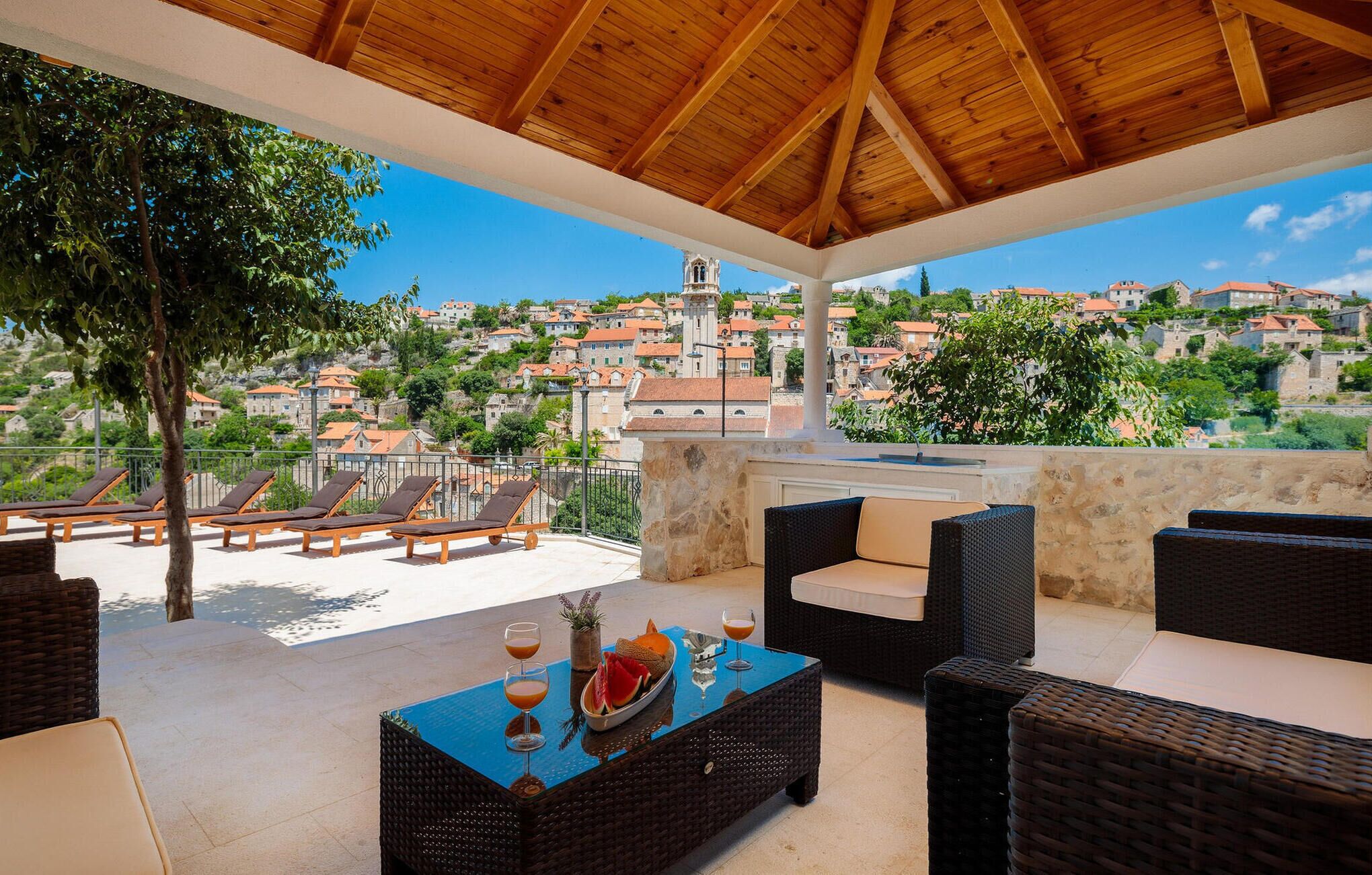 A covered outdoor seating area with wicker furniture, a table with drinks, and a view of a hillside town.